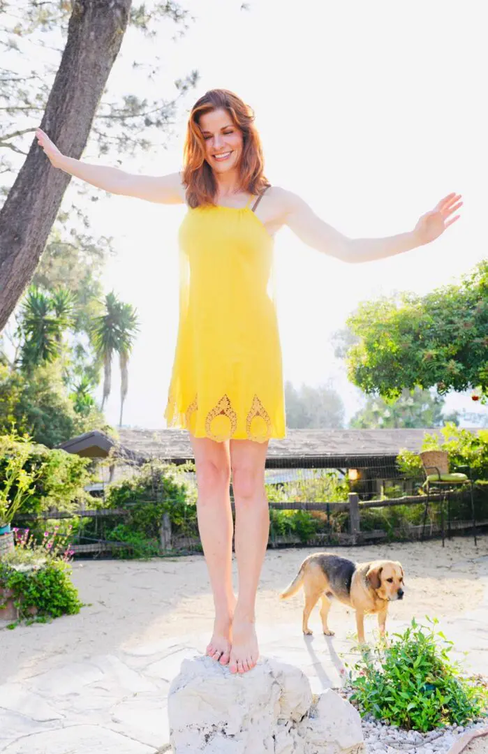 A woman in yellow dress standing on one foot with her arms outstretched.